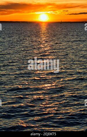 The sun sets over the Ottawa River at Lac Deschenes, with sailboats sailing in the distance. The sun's light reflects in a vertical pattern in this po Stock Photo