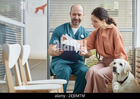 Portrait of young woman talking to smiling veterinarian in waiting room at vet clinic with white dog, copy space Stock Photo