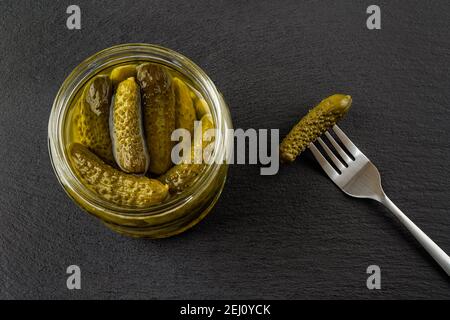 Open glass jar of tasty pickled gherkins and one more on the spoon against black slate background. Whole green cornichons marinated with dill. Stock Photo
