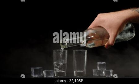 Bartender pouring up frozen vodka from bottle into two shots glasses with ice cubes isolated on black background. Barman pour of cold transparent alcohol drink vodka tequila. Copy-space Stock Photo