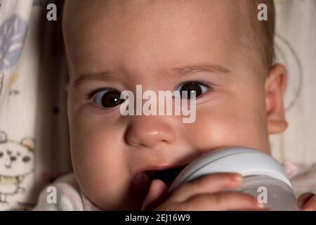 infant, childhood, emotion, food concept - close-up of smiling face of big brown-eyed chubby newborn awake baby 7 months drinks water from bottle Stock Photo