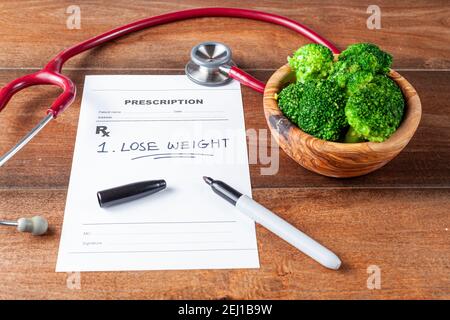 Close up angled image of a doctor's office desk with a bowl of broccoli,  stethoscope, pen and a prescription that has one item 'Lose weight' prescrib Stock Photo