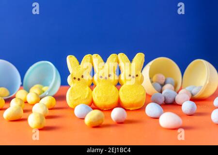 Easter holiday celebration concept with three little marshmallow bunnies and multiple small colorful crisp sugar coated chocolate easter eggs on orang Stock Photo