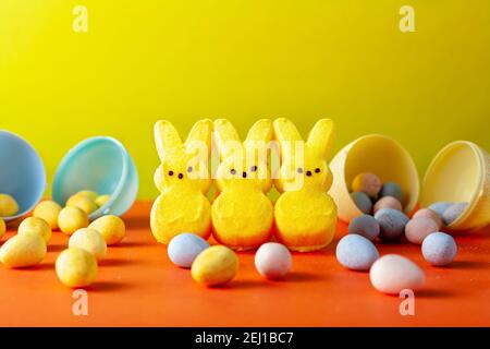 Easter holiday celebration concept with three little marshmallow bunnies and multiple small colorful crisp sugar coated chocolate easter eggs on orang Stock Photo
