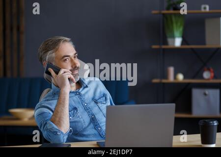 Senior grey-haired businessman talking on the phone in office. Conversation with business partners. Business portrait of handsome mature man sitting Stock Photo