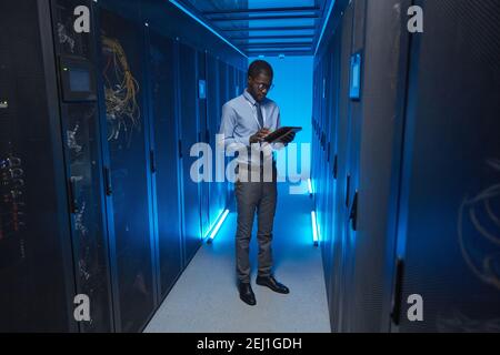 Full length portrait of African American man standing by server cabinet while working with supercomputer in data center and holding tablet, copy space Stock Photo