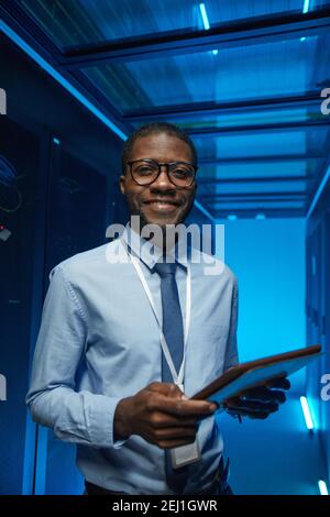 Vertical waist up portrait of smiling African American man standing by server cabinet while working with supercomputer in data center and holding tabl Stock Photo