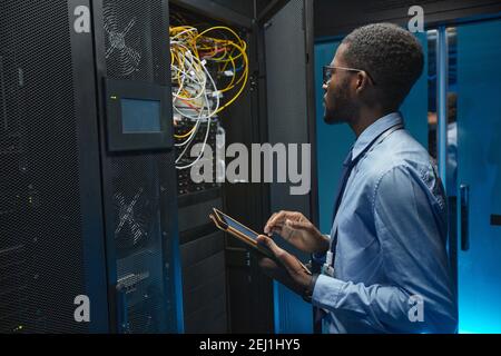 Side view portrait of African American man standing by server cabinet while working with supercomputer in data center and holding tablet, copy space Stock Photo