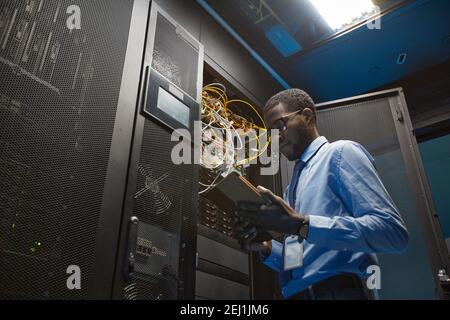 Low angle of African American man holding digital tablet while standing by server cabinet and working with supercomputer in data center, copy space Stock Photo