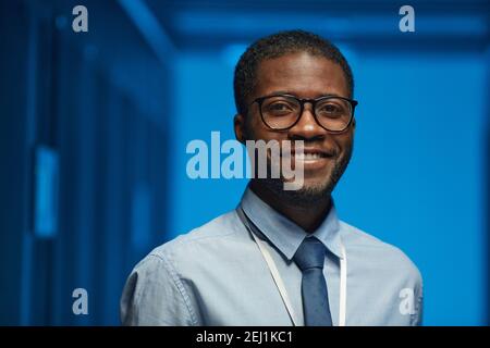 Portrait of smiling African American man wearing glasses and looking at camera while posing in server room at data center lit by blue light, copy spac Stock Photo
