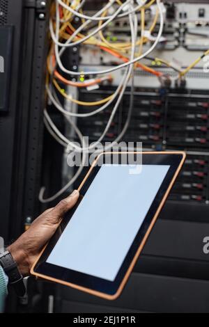 Vertical closeup of African American data engineer holding digital tablet with blank white screen while working with supercomputer in server room lit Stock Photo