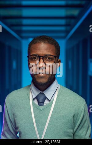 Vertical portrait of African American man wearing glasses and looking at camera while standing in server room with supercomputer at data center Stock Photo