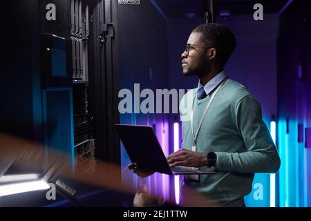 Side view portrait of African American data engineer holding laptop while working with supercomputer in server room lit by blue light, copy space Stock Photo