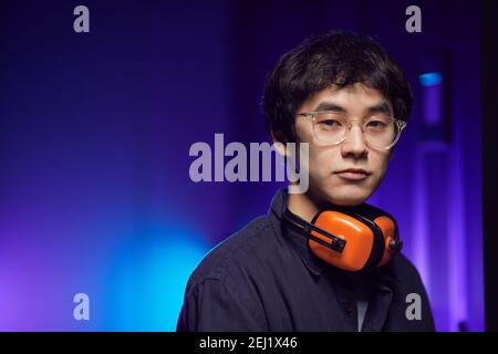Portrait of young Asian man wearing headphones looking at camera while standing in futuristic interior lit by blue light, copy space Stock Photo