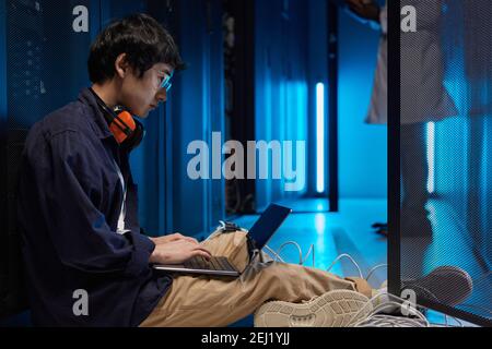Side view portrait of young Asian man sitting on floor in server room while setting up supercomputer network, copy space Stock Photo