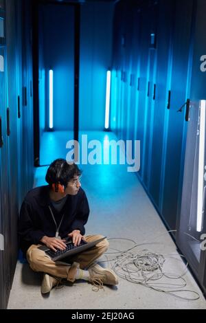 Vertical high angle portrait of young Asian man sitting on floor in server room lit by blue light while setting up supercomputer network via laptop, c Stock Photo