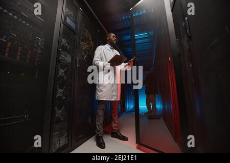 Full length portrait of African American data scientist wearing lab coat while working with supercomputer in server room, copy space Stock Photo