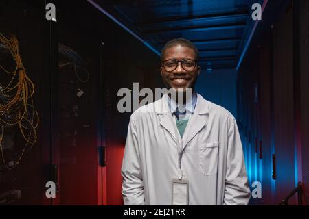 Waist up portrait of African American data scientist wearing lab coat and smiling at camera while working with supercomputer in server room, copy spac Stock Photo