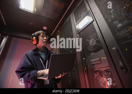 Waist up portrait of young Asian man using laptop while setting up supercomputer network in server room, copy space Stock Photo