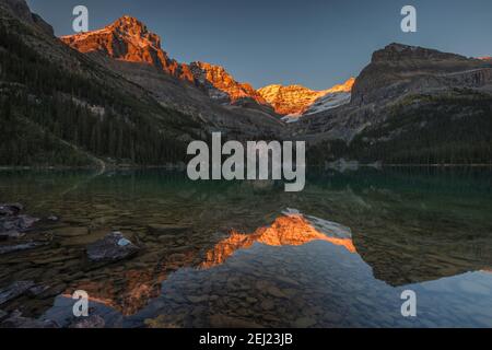 Sunset during fall in the mountains with snow, warm orange light on the peak, reflections on lake with calm clear water, trees and rocks, Canada Stock Photo