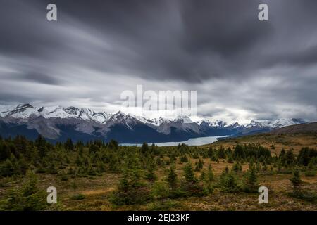 Long exposure of a Canadian Rockies landscape with a lake next to mountains with snow during autumn under a looming overcast sky with fast dark clouds Stock Photo