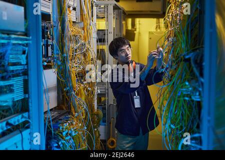 Wide angle portrait of young network technician connecting wires and cables in server room lit by blue light, copy space Stock Photo