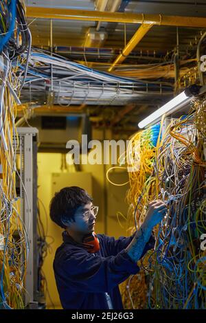 Vertical side view portrait of young network technician connecting wires and cables in server room, copy space Stock Photo