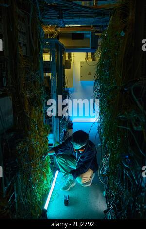Vertical high angle view at young network technician sitting on floor in server room and connecting cables, copy space Stock Photo