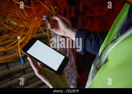 High angle close up of network technician holding smartphone with blank white screen in server room while inspecting equipment, copy space Stock Photo