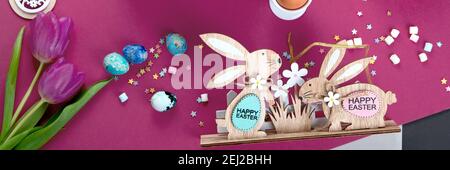 Happy Easter! Painted Easter eggs, decorative wooden bunnies and tulips. Dark/pink background. Top view, panorama banner Stock Photo