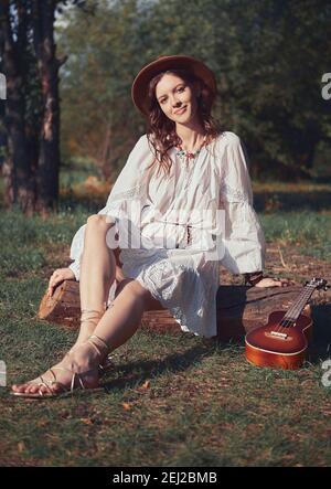 Outdoor portrait of the beautiful young boho (hippie) girl in glade (meadow). Pretty smiling woman with ukulele sits on stump at sunset time Stock Photo