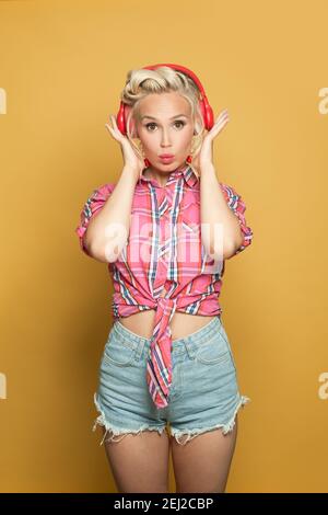 Pretty pinup woman listening music with headphones on bright yellow background. Pretty retro model with red lips makeup and vintage fashion hairstyle Stock Photo