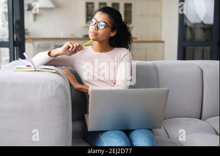 Thoughtful multiracial young woman with dark ponytail, sitting on the sofa with laptop and looking away, holding pencil in the hand near the face Stock Photo