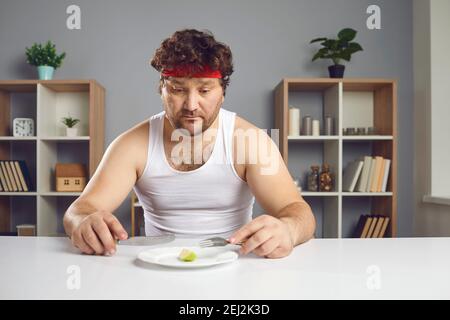 Sad man on strict diet sitting at table and looking at tiny piece of apple on plate Stock Photo