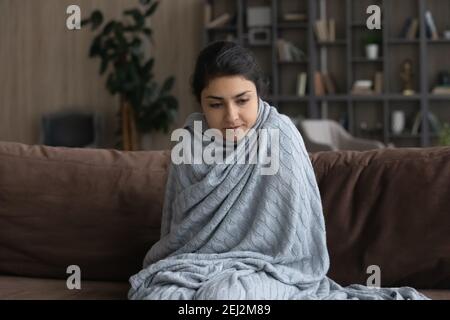 Sick young Indian woman wrapped in blanket feeling unhealthy