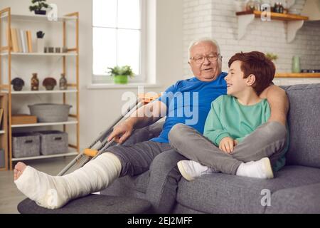 Happy grandson and his grandfather with broken leg in cast sitting on couch and talking Stock Photo