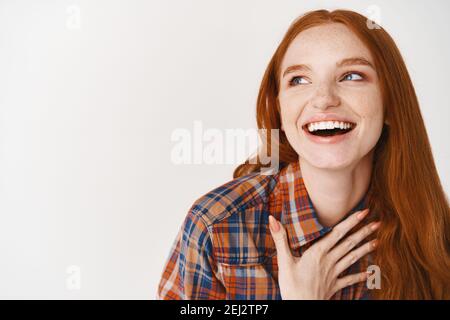 Close-up of beautiful woman with red hair and pale skin laughing happy, holding hand on heart and looking left at logo, standing over white background Stock Photo