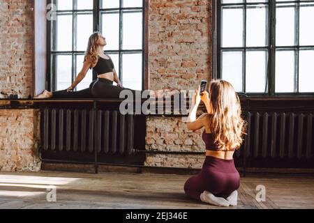 Two female friends take photo after hard workout loft style studio. Woman make picture of her flexible friend making splits on windowsill Stock Photo