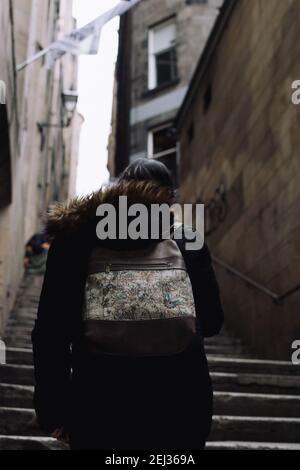 Young woman on her back wearing a black coat and a backpack going upstairs in an gloomy alley in Edinburgh Stock Photo