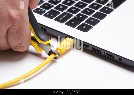 A closeup shot of a person cutting a yellow network cable with scissors connected to a laptop Stock Photo