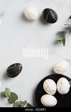 Elegant white and black Easter eggs decorated gold and eucalyptus leaves on marble table. Flat lay, top view, vertical. Stock Photo