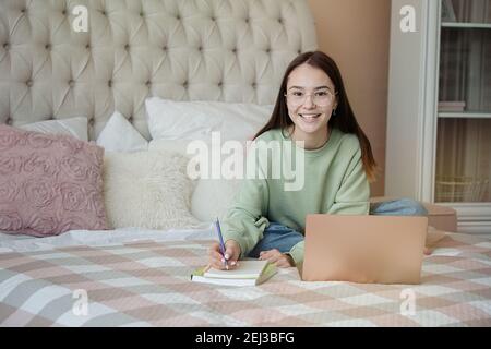 Cute teenager girl doing homework.Young girl wearing glasses writing down in notebook, studying online with laptop, distance learning, self education Stock Photo