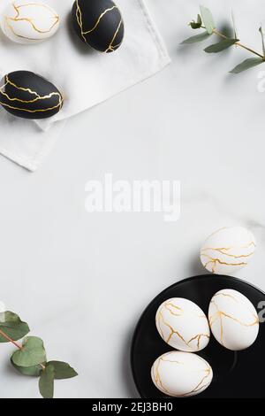 Frame of white and black Easter eggs decorated gold and eucalyptus leaves on marble table. Flat lay, top view. Happy Easter vertical banner design. Stock Photo