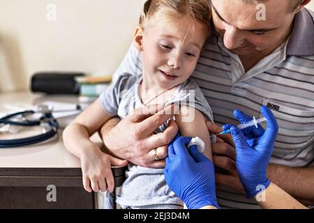 A little girl with her father in the doctor's office at the clinic is being vaccinated against the coronavirus. The concept of vaccination, immunizati Stock Photo