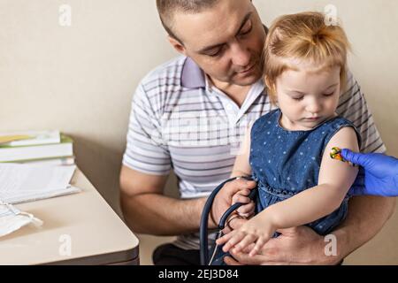Vaccination of a little girl in her dad's arms in the doctor's office from the coronavirus. Children's funny adhesive plaster. Vaccine against covid-1 Stock Photo