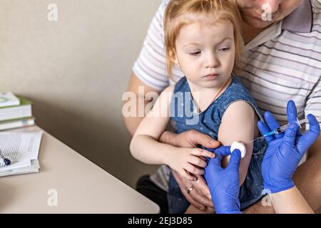 A little girl with her father in the doctor's office at the clinic is being vaccinated against the coronavirus.The concept of vaccination, immunizatio Stock Photo