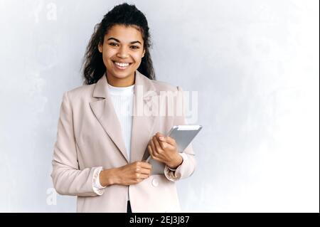 Portrait of a satisfied mixed race black young business woman or student in stylish formal wear, holds tablet, stands on isolated background, looks and smiles friendly at the camera Stock Photo