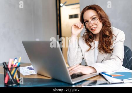 Stylish attractive young business lady sit at the desk, friendly smiling and looks at the camera. Redhead female employee using laptop, develop a new project, browsing internet Stock Photo