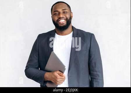 Portrait of an attractive African American male entrepreneur, freelancer or student in stylish suit, standing in the office holding laptop, looking and smiling friendly at the camera Stock Photo