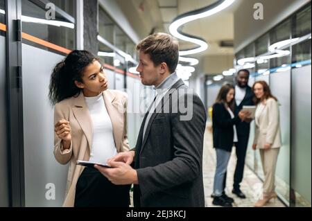 Two multiracial successful colleagues communicate in a modern office, discuss new ideas, think about a plan, in the background their colleagues Stock Photo